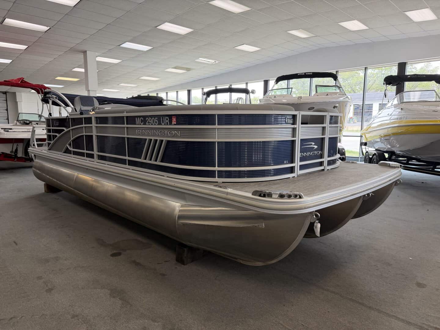 Pontoon boat inside showroom with large windows and other boats in the background.